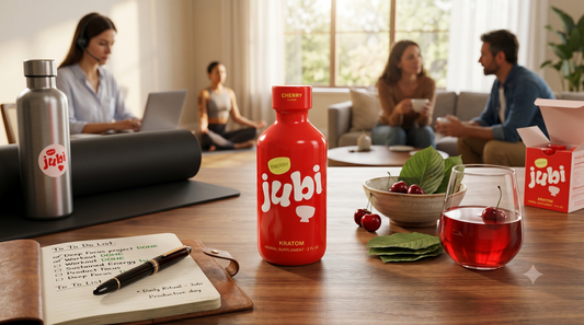 An advertisement for a wellness supplement featuring a small red bottle labeled "jubi" on a wooden table, surrounded by a journal, a glass of red drink, and fresh cherries, with people working and relaxing in the background.