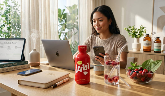  A young woman uses her phone at a bright, modern desk featuring a Jubi cherry energy shot, a laptop, a wellness journal, and fresh berries.