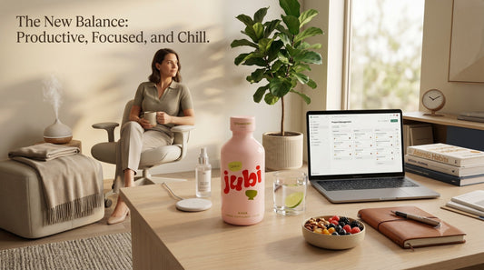 An advertisement featuring a bottle of Jubi Strawberry Kava on a modern wooden desk alongside a laptop, notebook, and healthy snacks, while a professional relaxes in the background.